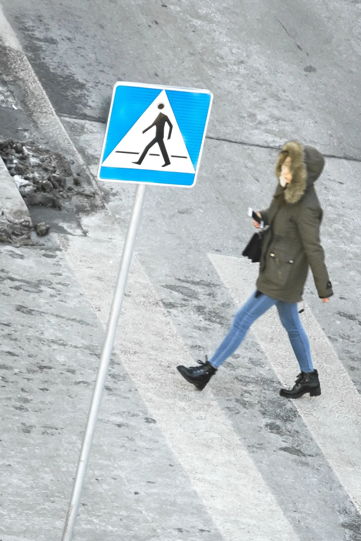 Overhead view of a bundled pedestrian walking past a blue crossing sign while holding a phone